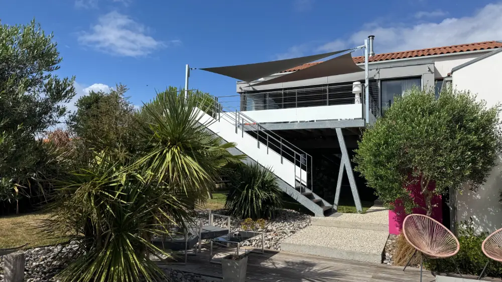 Deux voiles d’ombrage grises tendues au-dessus de la terrasse d’une maison, dans un jardin planté de palmiers et d’oliviers