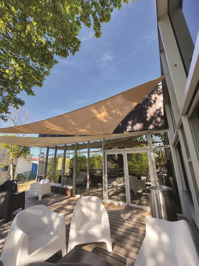 Voiles d’ombrage beiges tendues au-dessus d’une terrasse en bois avec fauteuils blancs, devant une façade vitrée